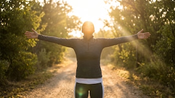 Una persona con ropa deportiva gris permanece de pie con los brazos extendidos y el rostro hacia el sol en un camino arbolado.