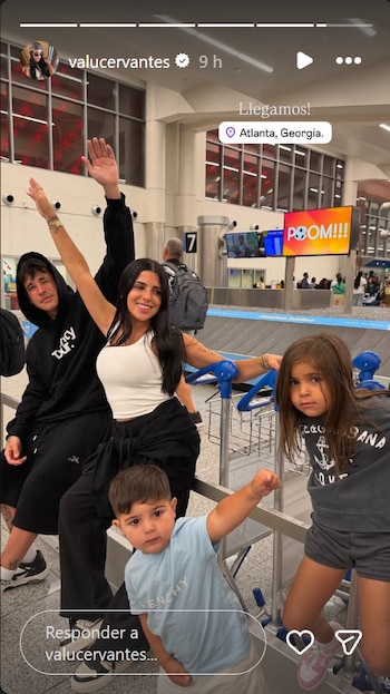Valentina Cervantes sonríe y levanta un brazo junto a un hombre y tres niños frente a una cinta de equipaje en un aeropuerto, con un cartel digital al fondo