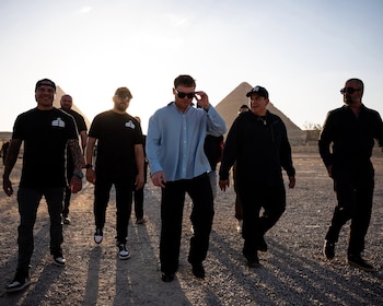 Grupo de hombres camina por un sendero de grava con las pirámides de Giza al fondo, bajo un cielo despejado. Un hombre de camisa azul ajusta sus gafas de sol