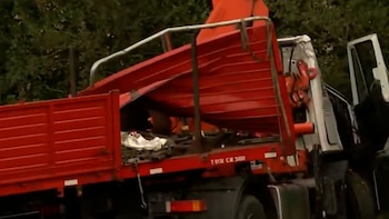 Vista lateral de un camión comercial blanco y rojo con la caja de carga elevada, transportando una estructura roja grande y piezas de maquinaria. Fondo de árboles verdes