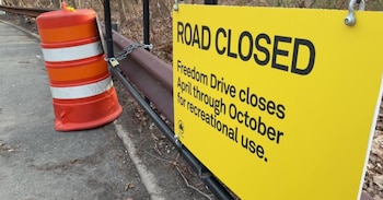 Un letrero amarillo de "ROAD CLOSED" y un barril de tráfico naranja bloquean una calle en Nueva York, con una barandilla metálica