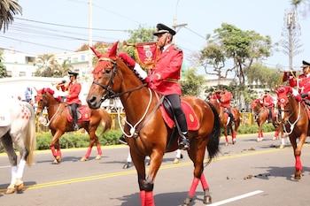 Jinetes militares con uniformes rojos desfilan a caballo en una calle. Los caballos son marrones y blancos, con adornos rojos. Algunos jinetes tocan trompetas