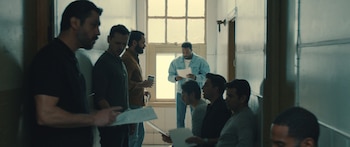Un grupo de hombres, incluido Yahya Abdul-Mateen II, se congrega en una sala con ventanas, algunos sentados, otros de pie, leyendo papeles