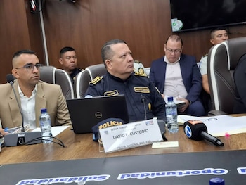 Hombres sentados en una mesa con micrófonos, botellas con agua, una computadora portátil y una placa con el nombre de un comisario de policía