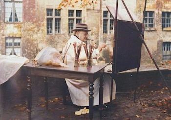 Winston Churchill, con sombrero y bata, pinta en un caballete al aire libre. Hay pinceles, un cigarro y un edificio de piedra de fondo