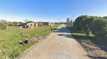 Calle de tierra con vegetación a los lados, postes de luz, alambrados y construcciones de ladrillo bajo un cielo despejado