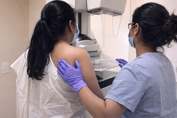 Dos mujeres de espaldas en una sala médica: una paciente con bata blanca frente a un mamógrafo, asistida por una profesional de la salud con guantes y mascarilla.
