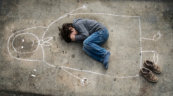 Un niño yace acurrucado sobre un pavimento gris con ropa oscura y jeans, cubriendo su rostro con los brazos, junto a un dibujo de tiza y un par de zapatos marrones.