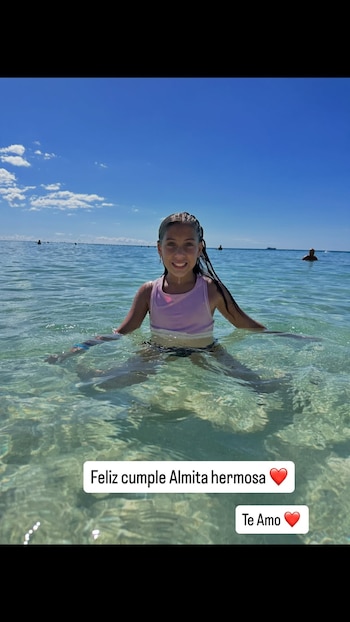 Una niña sonríe en el agua cristalina hasta la cintura, con traje de baño lila y negro, bajo un cielo azul con nubes. Hay texto flotante