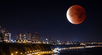 Una luna llena de tono rojo en un cielo nocturno sobre la costa e iluminada silueta urbana de la ciudad