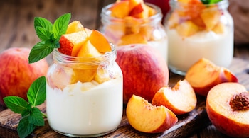 Frascos de vidrio con yogurt y trozos de durazno, junto a duraznos enteros y rebanados sobre una superficie de madera.