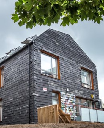 Casa de dos pisos revestida con tejas oscuras, ventanas con marcos de madera y una estructura de listones en la entrada
