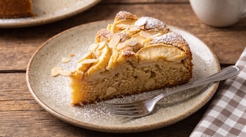 Porción de pastel de manzana decorada con almendras laminadas y azúcar impalpable sobre un plato con tenedor en una mesa de madera.