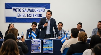 Hombre en traje habla por micrófono en un panel con banderas de El Salvador. Un banner azul y blanco dice 'VOTO SALVADOREÑO EN EL EXTERIOR'. Audiencia al frente.
