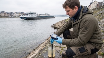 Un hombre en la orilla de un río sostiene un medidor digital sumergido en un vaso con agua, mientras una embarcación pasa al fondo.