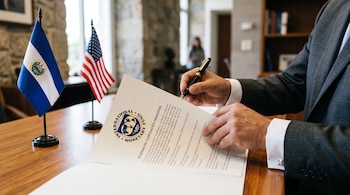 Manos sostienen una estilográfica sobre un documento del Fondo Monetario Internacional junto a banderas de El Salvador y Estados Unidos.