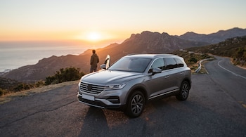 Un SUV plateado estacionado en una carretera asfaltada junto a un hombre que mira el atardecer entre montañas y el mar.