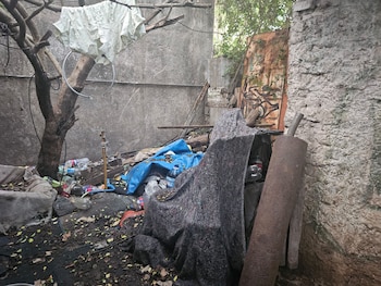 Un área exterior descuidada con un árbol, muros de concreto y piedra, ropa colgando y un suelo cubierto de basura, botellas y una lona azul