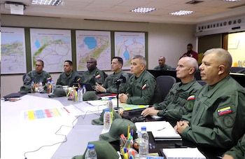 Ocho hombres en uniforme militar sentados en una sala de reuniones. Sobre la mesa, gorras, botellas de agua, cuadernos y bolígrafos. Tres mapas en la pared
