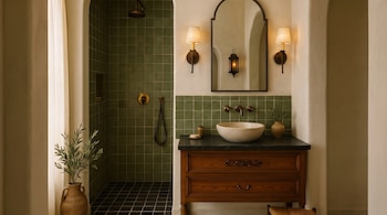 Interior de un baño con ducha de azulejos verdes, grifos de metal, lavabo de piedra, mueble de tocador de madera oscura, espejo arqueado, apliques de pared y una planta en maceta de arcilla.