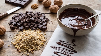 Barra de chocolate, trozos de chocolate, nueces enteras, nueces molidas, cuenco con chocolate derretido, cuchara de metal, cuchillo, papel de horno y mesa de madera.