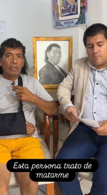 Cachay, con un cabestrillo negro en el brazo izquierdo, se sienta y habla, con un hombre a su lado que sostiene papeles. Al fondo, un retrato enmarcado