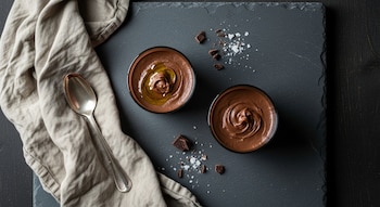 Vista cenital de dos copas de mousse de chocolate. Una con aceite de oliva y sal marina, con virutas de chocolate, cuchara y servilleta sobre tabla oscura.
