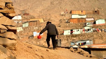 Una persona de espaldas con sombrero camina por un cerro de tierra con un balde y una jarra, frente a viviendas precarias