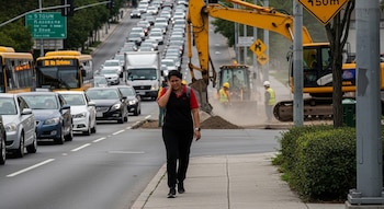 Mujer en primer plano camina en acera junto a calle con tráfico vehicular, dos autobuses, una excavadora, un bulldozer y trabajadores.