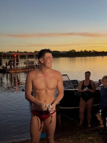Andy Donaldson, torso desnudo y bañador rojo, sonríe de pie en la orilla de un lago al atardecer, con un bote y personas al fondo