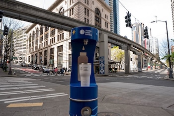 Una estación de agua azul de Seattle Public Utilities con una botella blanca debajo, ubicada en una acera de Downtown Seattle con un monorraíl y edificios de fondo
