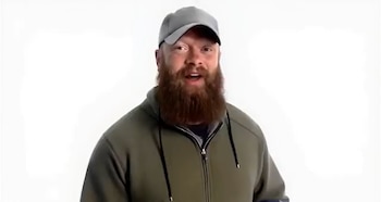 Retrato de un hombre barbudo con una gorra gris y una chaqueta deportiva verde frente a un fondo blanco