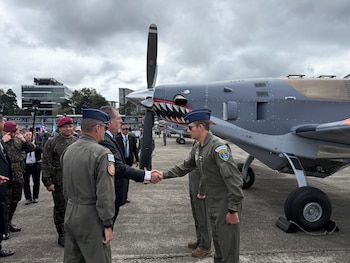 Dos militares con uniforme verde y gorro de cuartel azul están de pie junto a un avión de hélice decorado bajo un cielo nublado
