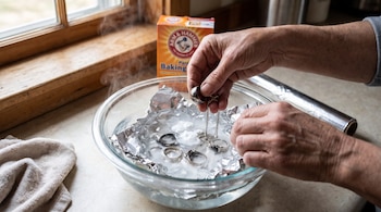 Manos de una persona limpiando anillos y cadena de plata en un cuenco con agua caliente, bicarbonato de sodio y papel aluminio, con una caja de bicarbonato.