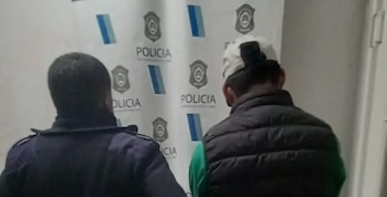 Vista trasera de dos hombres: uno con uniforme policial y otro joven con gorra, camiseta verde y chaleco oscuro. Al fondo, una pared con logos de la Policía
