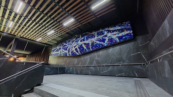 Vista interior de la terminal Tasqueña del Metro CDMX, mostrando una gran obra de arte en mosaico azul sobre una pared de mármol oscuro, escaleras y un techo de listones de madera