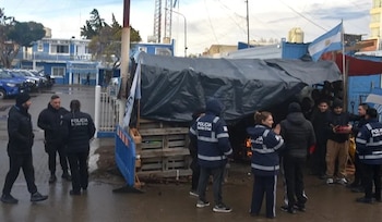 Edificio azul de Policía Santa Cruz, vehículos policiales, grupo de policías y civiles, refugio precario con lona negra, fuego, bandera argentina