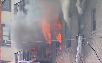 Edificio de ladrillo con ventanas en llamas, humo denso, balcón con barandilla y poste de servicios con cables