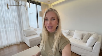 Mujer rubia con top verde oliva, de pie en una sala de estar moderna y luminosa con sofás blancos, un candelabro dorado y cortinas blancas sobre grandes ventanales.