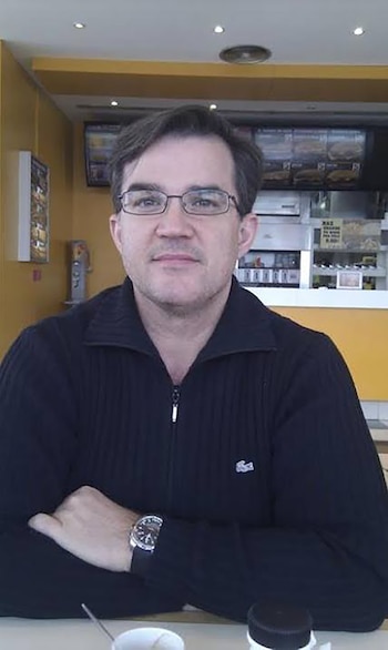 Hombre sentado con gafas, suéter negro con cremallera, brazos cruzados, y un reloj en la muñeca izquierda. Hay una mesa y un fondo amarillo