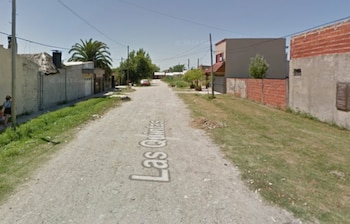 Calle de tierra "Las Quintas" en Berisso con casas a los lados, vegetación, postes de luz y una persona caminando a la izquierda bajo cielo azul claro