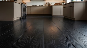 Primer plano de un piso de madera oscura con vetas visibles que se extiende hacia una cocina moderna con gabinetes beige, encimeras claras y un horno de acero inoxidable.