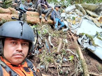 Primer plano de un rescatista con casco y cámara en la cabeza. Detrás hay un tronco de árbol, personas sentadas y restos de un avión en el suelo