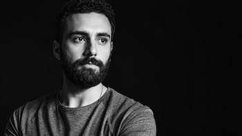 Fotografía en blanco y negro de un hombre con barba, bigote y cabello corto oscuro que mira hacia un costado sobre un fondo negro