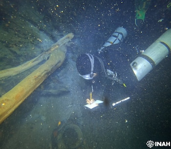 Buceador con máscara y equipo de buceo, escribiendo en libreta bajo el agua con lápiz. Flotan tanques de buceo y restos de madera en agua oscura