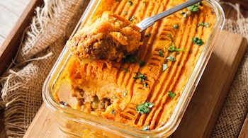 Recipiente de vidrio con pastel de zanahoria y carne. El pastel tiene una capa superior de puré anaranjado y se está sirviendo con una cuchara metálica.