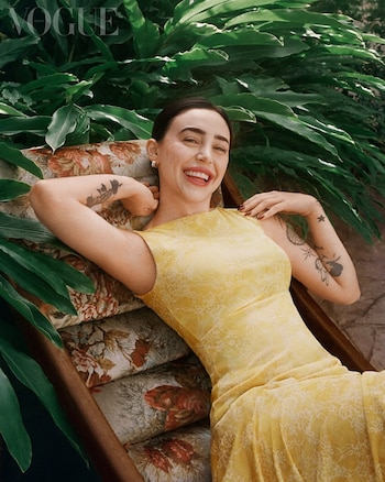 Nicki Nicole sonríe recostada en un sofá con cojines florales, vistiendo un vestido amarillo claro con encaje, rodeada de frondosas plantas verdes