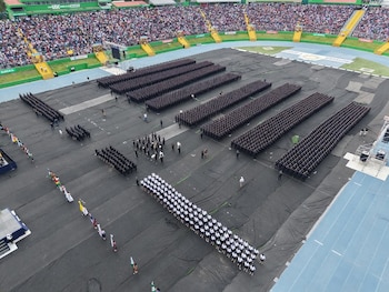 Vista desde las alturas de formaciones de uniformados en la cancha cubierta de un estadio lleno de personas