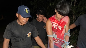 Tres hombres; uno con gorra azul y camiseta negra, otro con camiseta roja sin mangas y brazos atados con cuerda azul, y un tercero detrás.