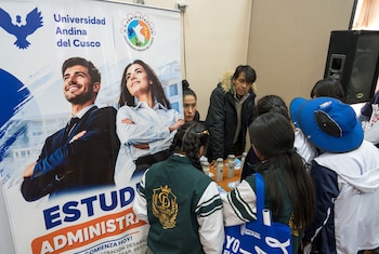 Estudiantes de espaldas frente a un puesto informativo con un cartel de la Universidad Andina del Cusco y dos personas detrás de una mesa